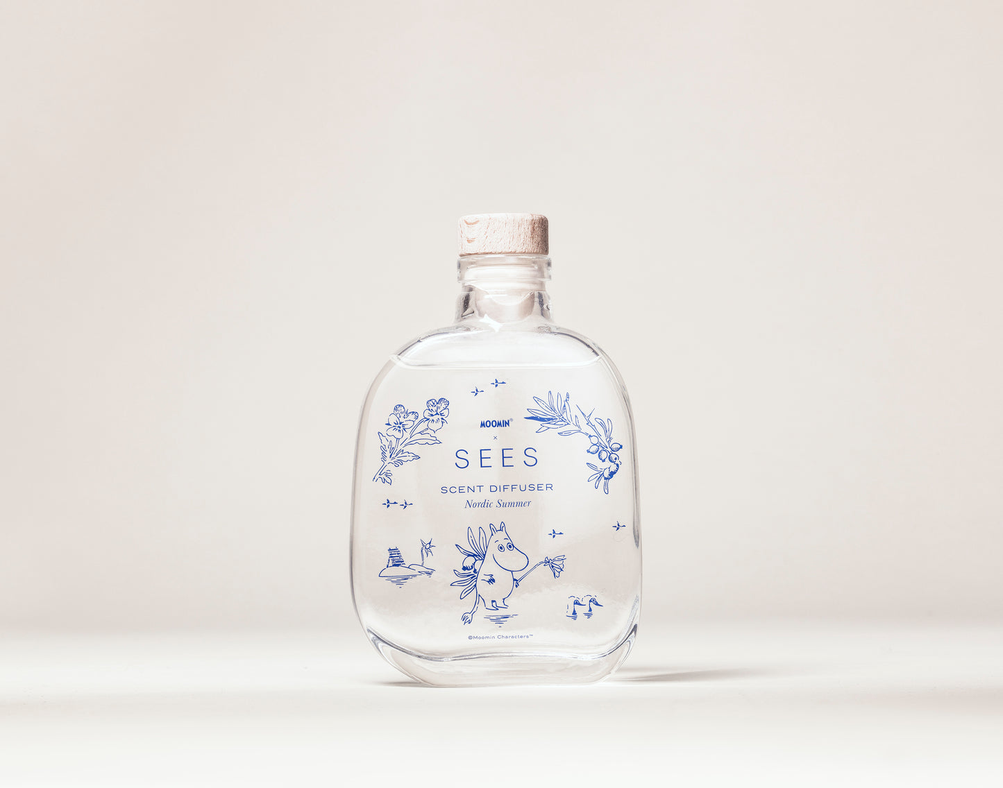 Eine klare Glasflasche mit Korkverschluss und der Aufschrift „SEES x Moomin Room Diffuser Nordic Summer by SEES Company“ mit blauen Blumenmustern, Blättern und einer kleinen Moomin-Figur steht auf einem schlichten hellen Hintergrund.