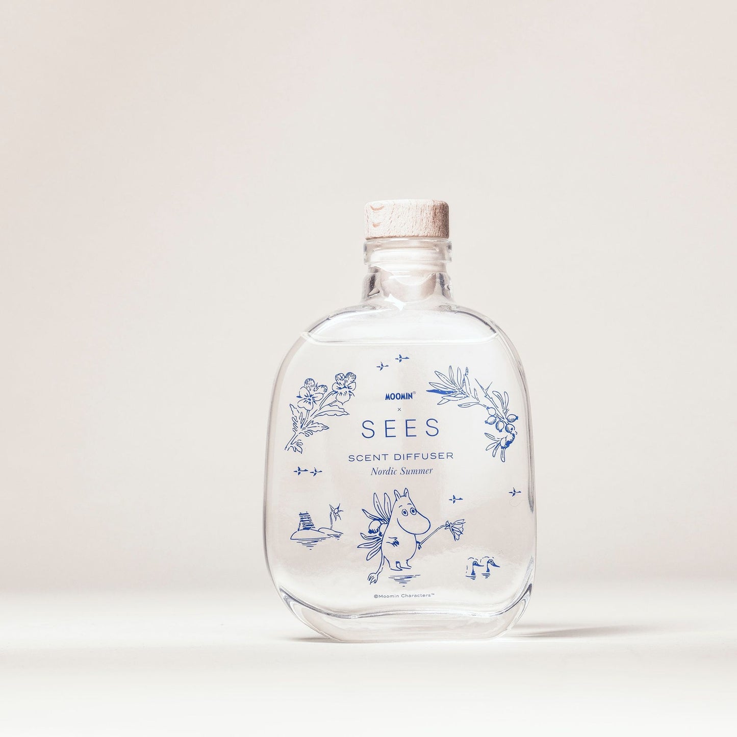 Eine klare Glasflasche mit Korkverschluss und der Aufschrift „SEES x Moomin Room Diffuser Nordic Summer by SEES Company“ mit blauen Blumenmustern, Blättern und einer kleinen Moomin-Figur steht auf einem schlichten hellen Hintergrund.