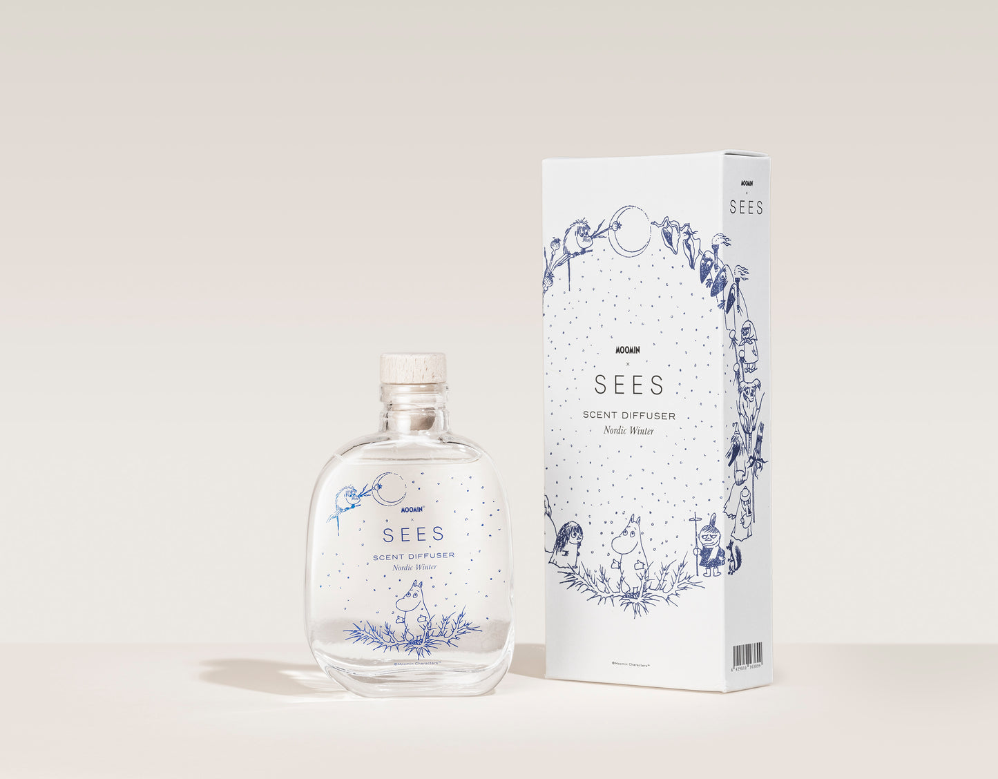 Ein klarer Glasdiffusor SEES x Moomin Room Diffuser Nordic Winter von SEES Company mit Holzverschluss steht neben einer weißen Schachtel mit verspielten blauen Illustrationen – eine Hommage an finnisches Design und die gemütliche Atmosphäre nordischer Winter.