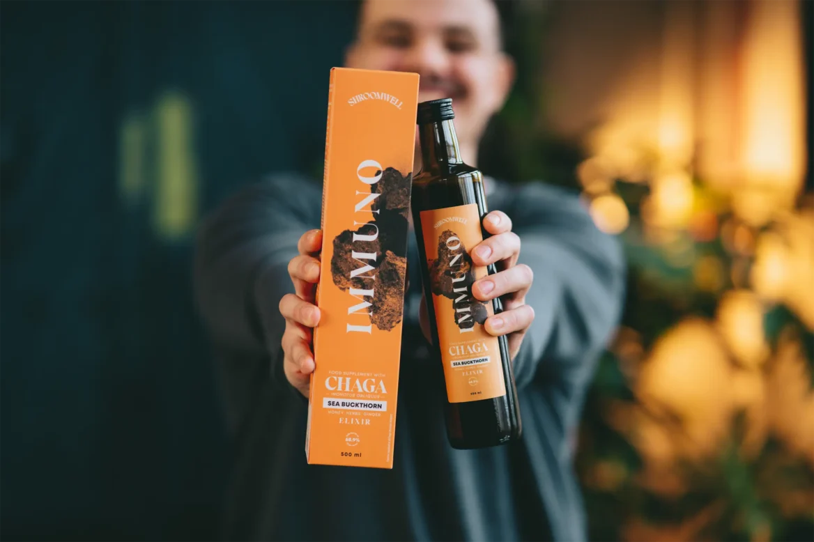 Eine Person zeigt das Shroomwell Immuno Elixir Chaga & Sanddorn, das seine wirksame Mischung aus Chaga-Pilz und Sanddorn zur Unterstützung des Immunsystems hervorhebt, vor einem sanft verschwommenen Hintergrund mit warmem Licht und Grün.