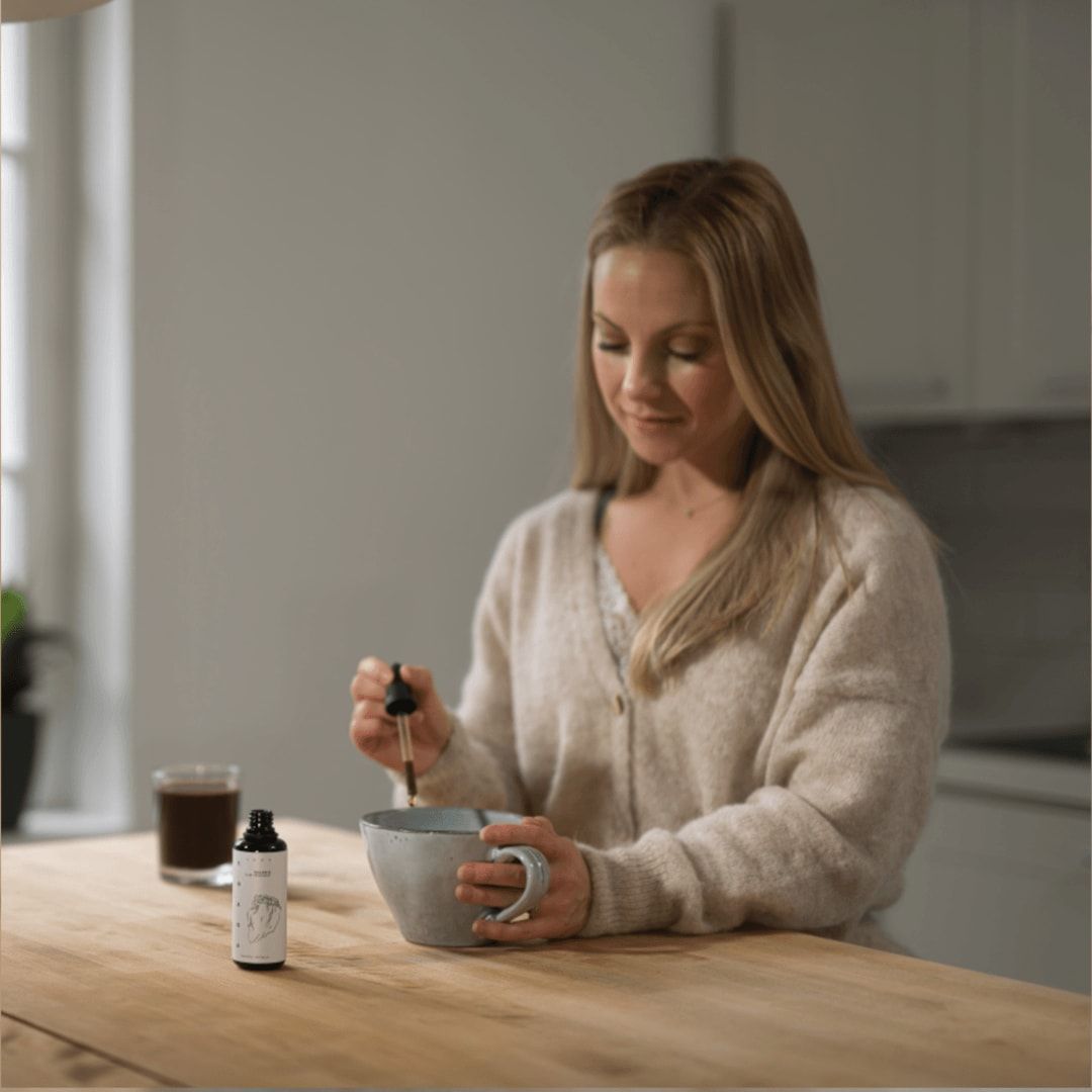 Eine Frau sitzt an einem minimalistischen Küchentisch und rührt ein Getränk mit einer Pipette um, die mit KÄÄPÄ Mushrooms Organic Chaga Extract gefüllt ist. Eine Flasche KÄÄPÄ Mushrooms Chaga und ein Glas mit dunkler Flüssigkeit stehen ebenfalls auf dem sanft beleuchteten Tisch.