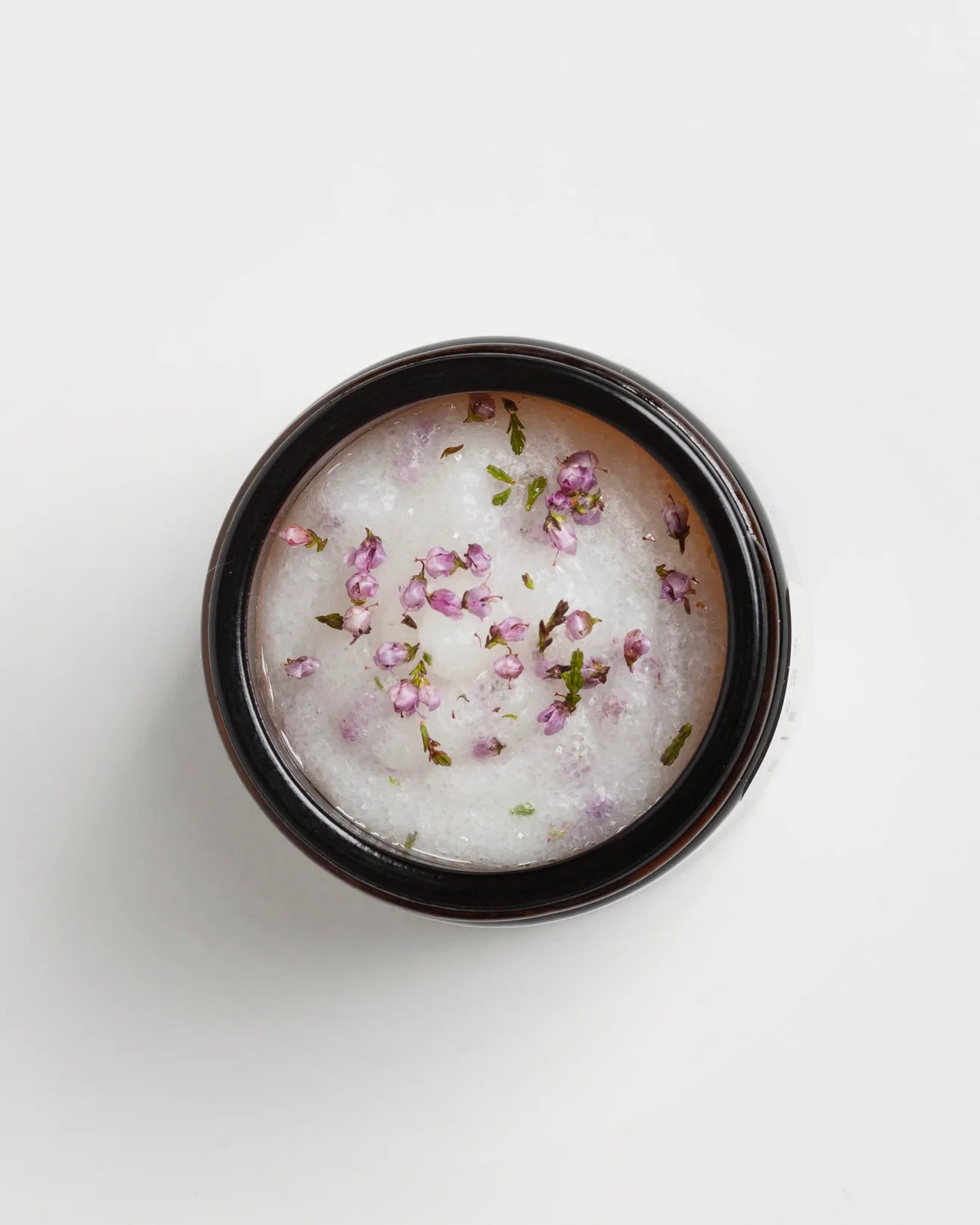 Das Hetkinen Metsä Calluna Scrub von Hetkinen ist ein cremiges Peeling für Körper und Gesicht in einem schwarzen Behälter mit violetten und grünen Blütenblättern, die von finnischen Waldblumen inspiriert sind, und einer weißen Oberfläche.