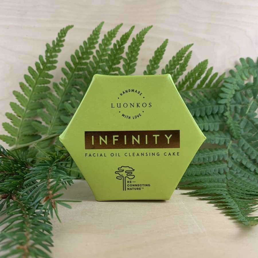 Eine sechseckige grüne Schachtel mit Luonkos Infinity Oil Cleansing Cake steht aufrecht auf einer hellen Fläche, umgeben von grünem Farn und Kiefernzweigen - ideal für empfindliche Haut.