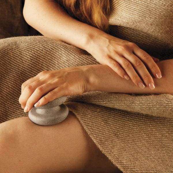Eine in ein braunes, strukturiertes Tuch gehüllte Person legt die Hukka Sauna-Joy Massagehilfe von Hukka auf ihren Oberschenkel, während die andere Hand sanft auf ihrem Schoß ruht.