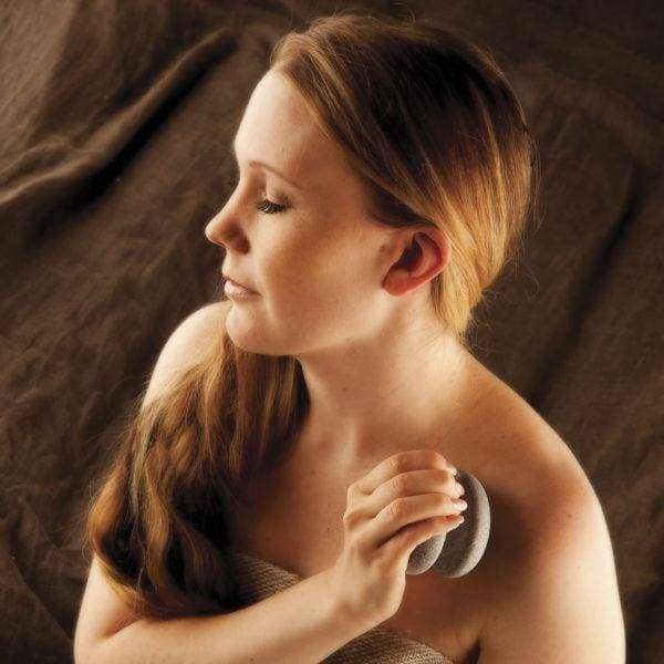 Eine Frau mit langen braunen Haaren, die in ein Handtuch gewickelt ist, peelt sanft ihre Schulter mit der Hukka Sauna-Joy Massagehilfe von Hukka vor einem weichen braunen Stoffhintergrund und schafft so einen beruhigenden Moment der Selbstpflege.