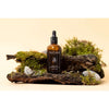 Eine braune Glastropfflasche mit Moi Forest Dust Microbiome Magic Oil von Moi Forest ist auf einer Rinde mit Moos und Steinen vor einem beigen Hintergrund abgebildet.