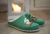 Une paire de Zapatillas végétaliens verts Zapatillas Felt Slippers Snufkin au motif cartoon repose sur un tapis, tandis qu'une bougie brille dans une lanterne derrière eux, créant une atmosphère chaleureuse.