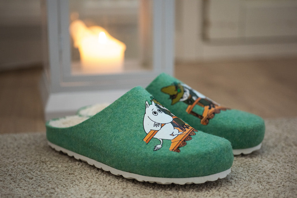 Une paire de Zapatillas végétaliens verts Zapatillas Felt Slippers Snufkin au motif cartoon repose sur un tapis, tandis qu'une bougie brille dans une lanterne derrière eux, créant une atmosphère chaleureuse.