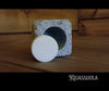 Une pastille ronde de sel blanc pour sauna provenant du pack de démarrage Kiuassuola Sauna Salt ILMARI repose devant un support carré en pierre avec un creux circulaire sur du bois, capturant l'essence même du bien-être. Le nom de la marque « Kiuassuola » apparaît en bas à droite.