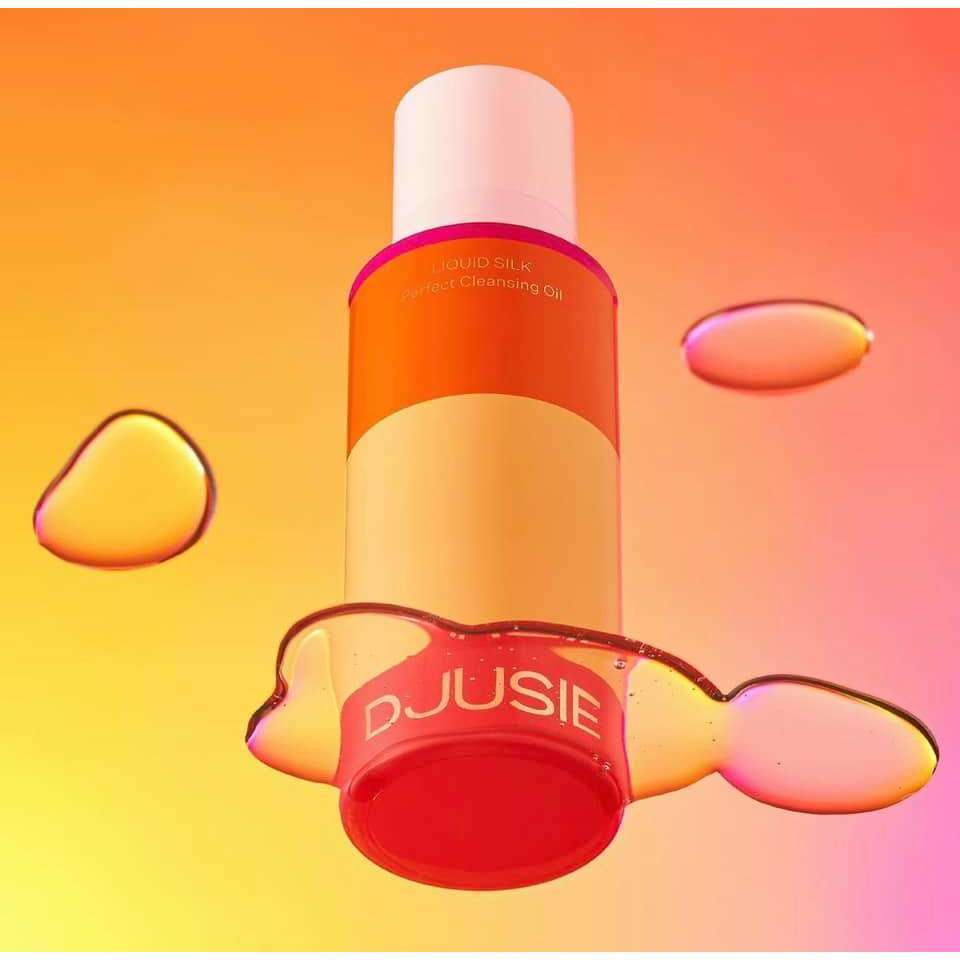 Une bouteille orange et jaune vif de l'huile démaquillante Djusie Liquid Silk Perfect Cleansing Oil semble flotter au-dessus d'un fond rose pêche, entourée de gouttelettes scintillantes. Idéale pour éliminer le maquillage waterproof.