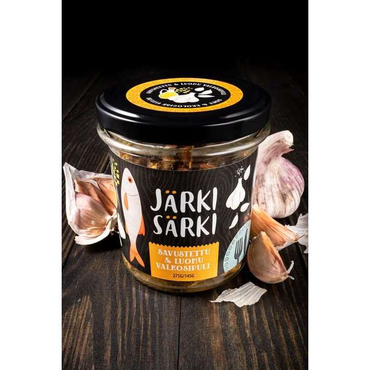 Un bocal en verre étiqueté « Järki Särki Smoked with Organic Garlic » (Järki Särki fumé à l'ail biologique) de la marque Järki Särki, orné d'illustrations représentant du poisson et de l'ail, repose sur une surface en bois foncé entourée de gousses d'ail frais. Cette spécialité nordique est préparée à partir de poisson sauvage pêché de manière durable.