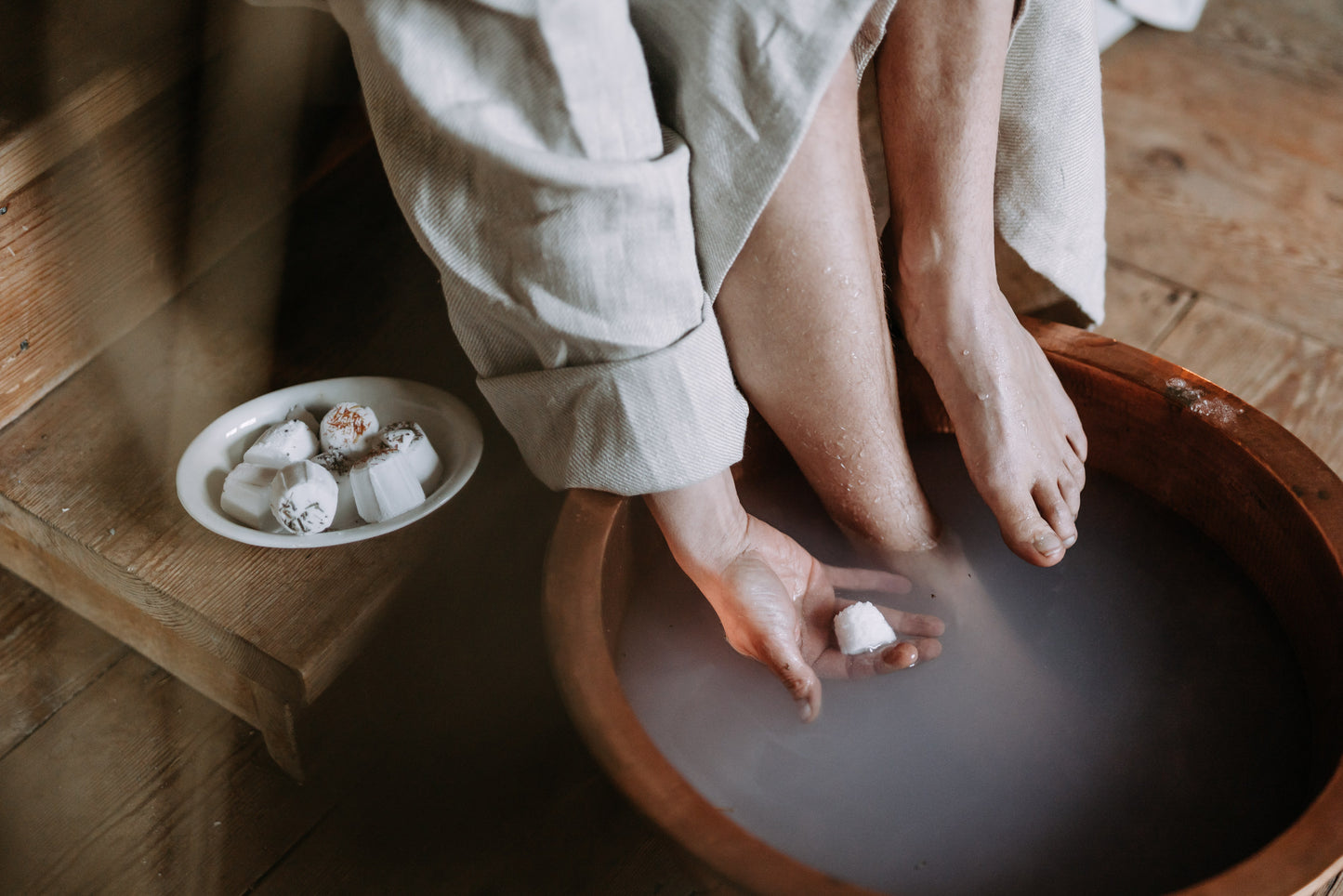 Une personne vêtue d'un peignoir trempe ses pieds dans une eau trouble à l'aide du bain de Kaurilan Sauna  Kaurilan Sauna, tandis qu'une assiette de bombes de bain cosmétiques naturelles repose sur les marches en bois à proximité.