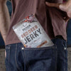 Une personne vêtue d'un jean bleu et d'une chemise marron a un paquet de Renjer Nordic Red Deer Jerky Chili & Lime de Renjer qui dépasse de sa poche arrière.