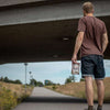 Un homme vêtu d'un t-shirt marron et d'un short en jean marche sur un chemin sous un pont, tenant un sac de Renjer Nordic Red Deer Jerky Chili & Lime, avec des champs verdoyants et un lampadaire en arrière-plan.