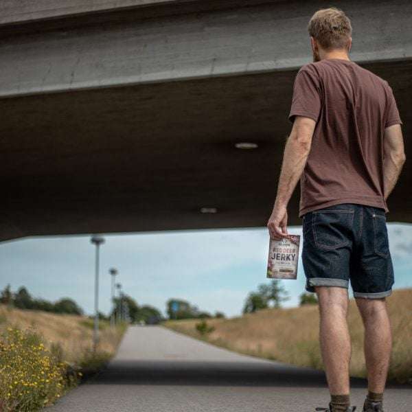 Un homme vêtu d'un t-shirt marron et d'un short en jean marche sur un chemin sous un pont, tenant un sac de Renjer Nordic Red Deer Jerky Chili & Lime, avec des champs verdoyants et un lampadaire en arrière-plan.