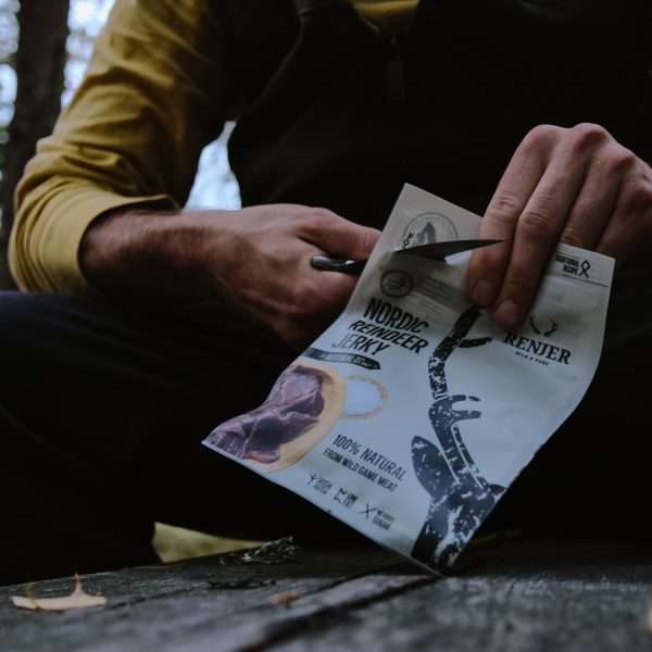 En plein air, dans un cadre rustique, quelqu'un ouvre un paquet de Renjer Nordic Reindeer Jerky Sea Salt de Renjer à l'aide d'un petit couteau, capturant ainsi les saveurs sauvages et les traditions nordiques du jerky de renne dégusté dans la nature.