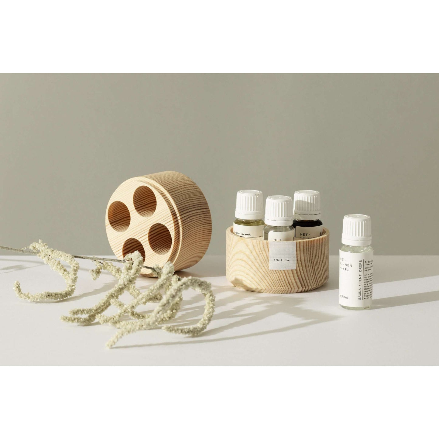 Quatre petites bouteilles Hetkinen Sauna Scent Woody avec des bouchons blancs et des étiquettes minimalistes sont posées dans un support rond en bois, évoquant un sauna finlandais. À proximité se trouve un morceau de bois avec des trous circulaires et des fleurs blanches séchées sur une surface claire, sur un fond neutre.