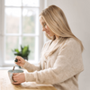 Une femme aux longs cheveux blonds sourit en ajoutant de l'extrait KÄÄPÄ Mushrooms de crinière de lion KÄÄPÄ Mushrooms dans sa tasse, assise à une table en bois, soucieuse de la santé de son cerveau. Une grande fenêtre et une plante en pot créent une atmosphère paisible et doucement éclairée.