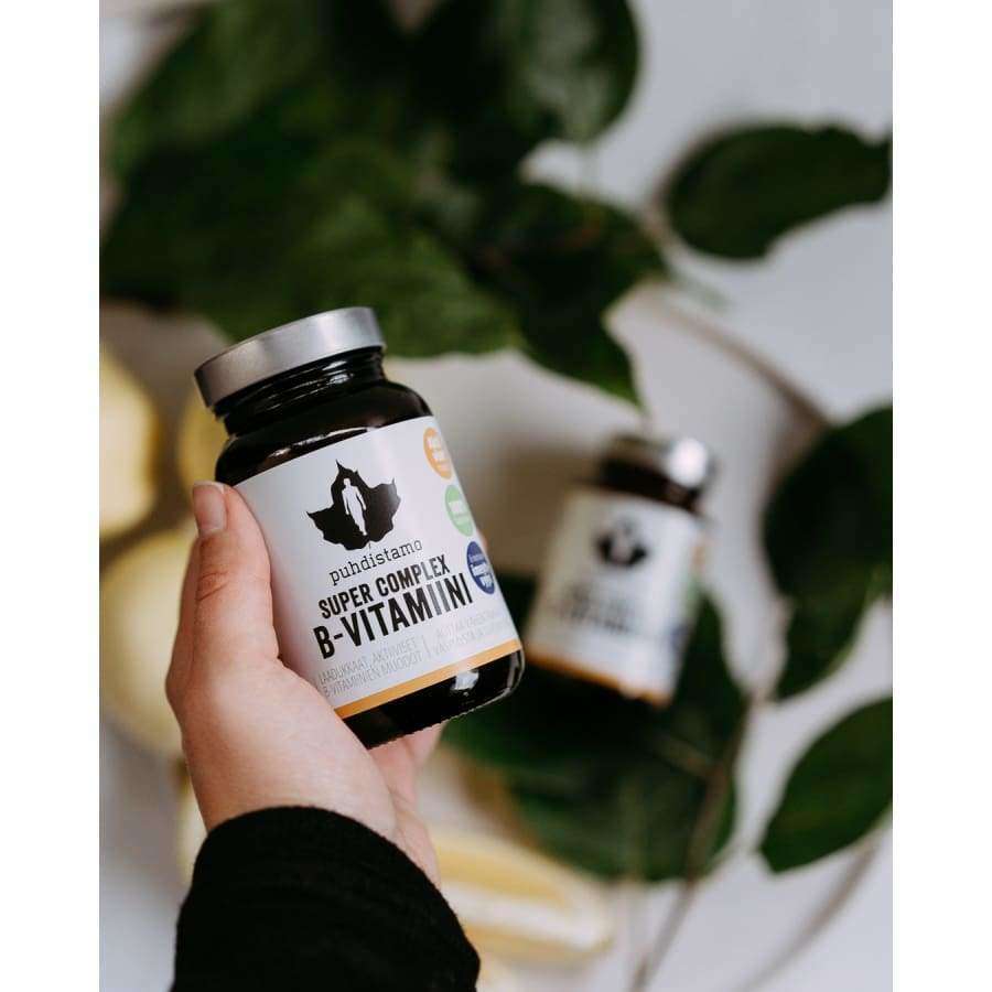 Une main tient un flacon de Puhdistamo Super Complex B-complex, un complément alimentaire à base de vitamine B hautement assimilable de Puhdistamo, avec un autre flacon et des feuilles vertes floues en arrière-plan.