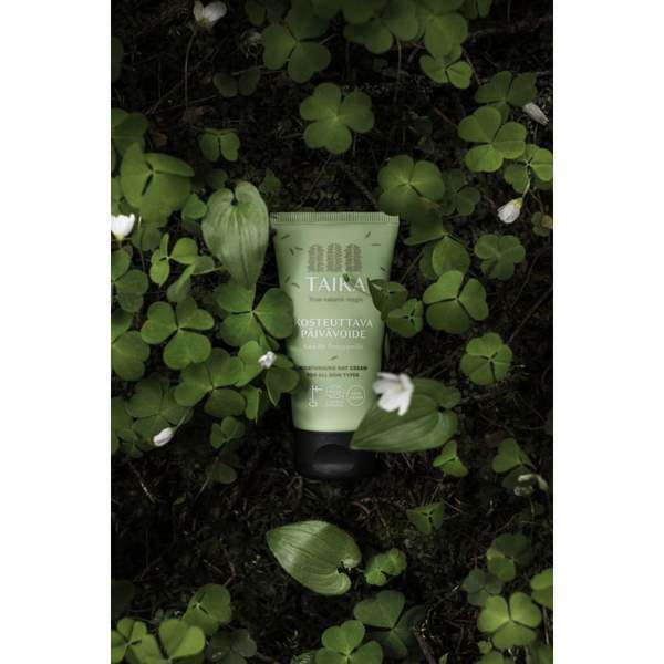 Un tube vert de crème de jour Taika repose parmi des feuilles de trèfle et de petites fleurs blanches, soulignant sa formule vegan pour les soins de la peau.