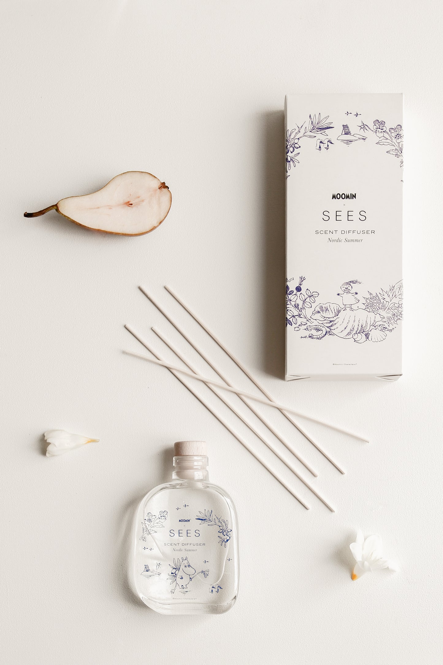 Una composizione piatta del diffusore SEES x Moomin Room Diffuser Nordic Summer di SEES Company, che mostra una scatola decorativa, una bottiglia di vetro etichettata, quattro bastoncini diffusori, mezza pera e petali di fiori bianchi su uno sfondo bianco.
