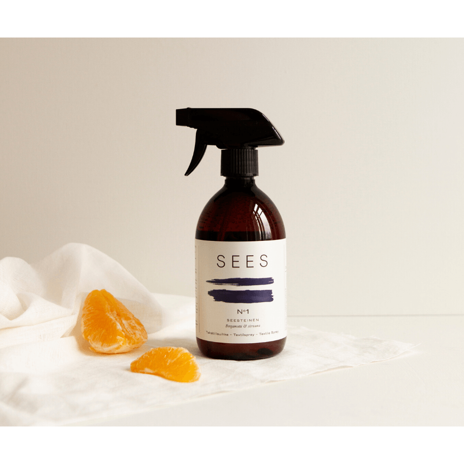 Una bottiglia marrone SEES Company Spray No. 1 Serene, Bergamot & Lemon poggia su un panno bianco accanto a due spicchi di mandarino arancione, con uno sfondo bianco e un'illuminazione soffusa.
