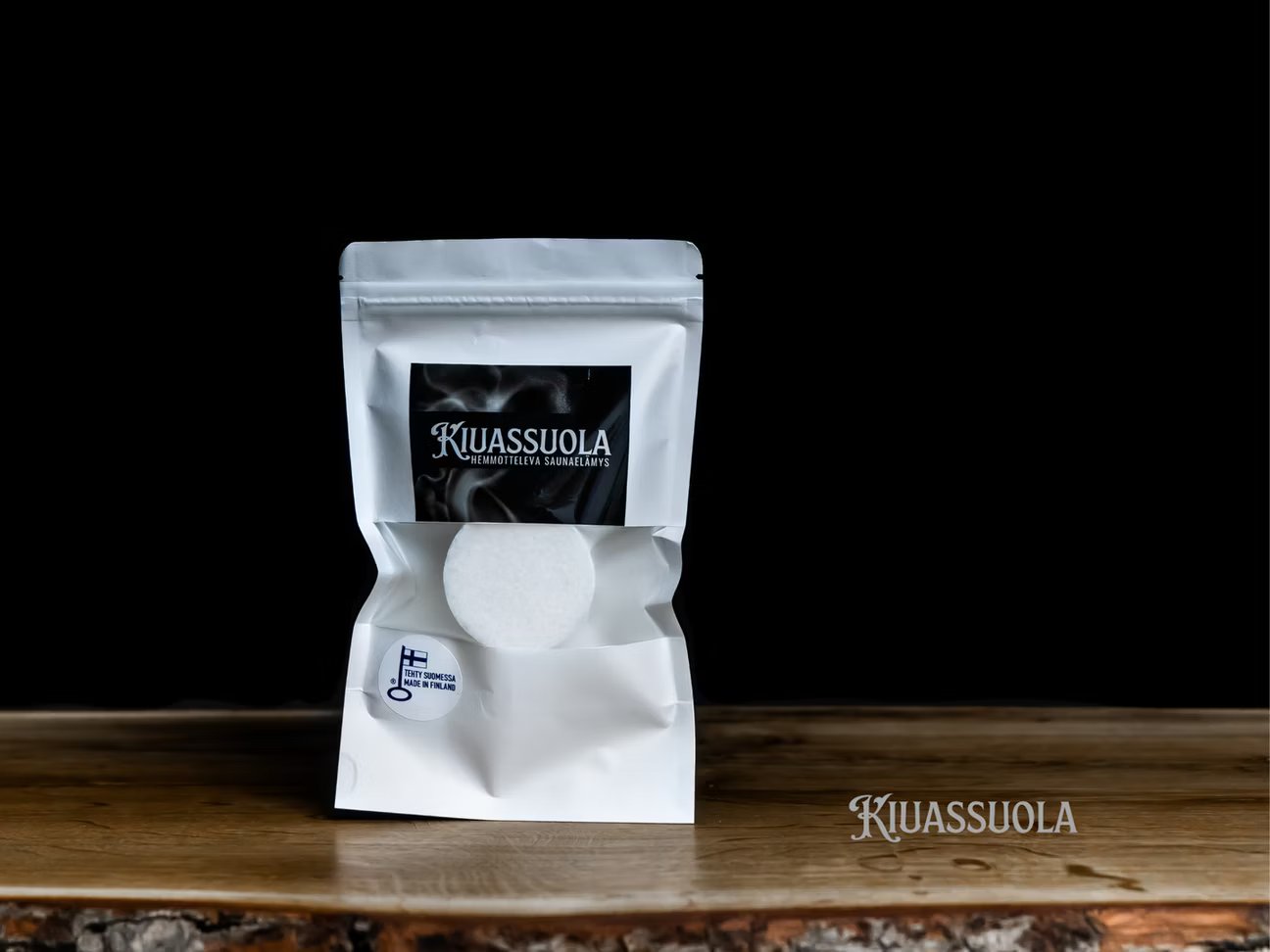 Una busta bianca richiudibile di sale per sauna Kiuassuola è appoggiata su una superficie di legno su uno sfondo nero. Davanti è posizionata una pastiglia rotonda di sale per sauna bianca, con il logo Kiuassuola visibile nell'angolo in basso a destra.