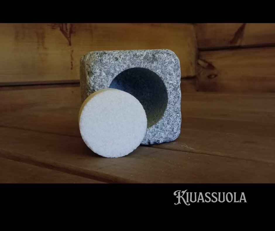 Una pastiglia rotonda di sale bianco per sauna della confezione iniziale Kiuassuola Sauna Salt ILMARI è posizionata davanti a un supporto quadrato in pietra con una cavità circolare su legno, che cattura l'essenza del benessere. Il marchio "Kiuassuola" appare in basso a destra.