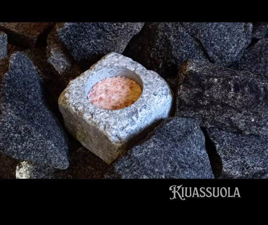 Il pacchetto iniziale Kiuassuola Sauna Salt ILMARI contiene un blocco di sale naturale rosato per sauna in un contenitore quadrato in pietra, circondato da pietre scure: perfetto per migliorare il tuo rituale di benessere.