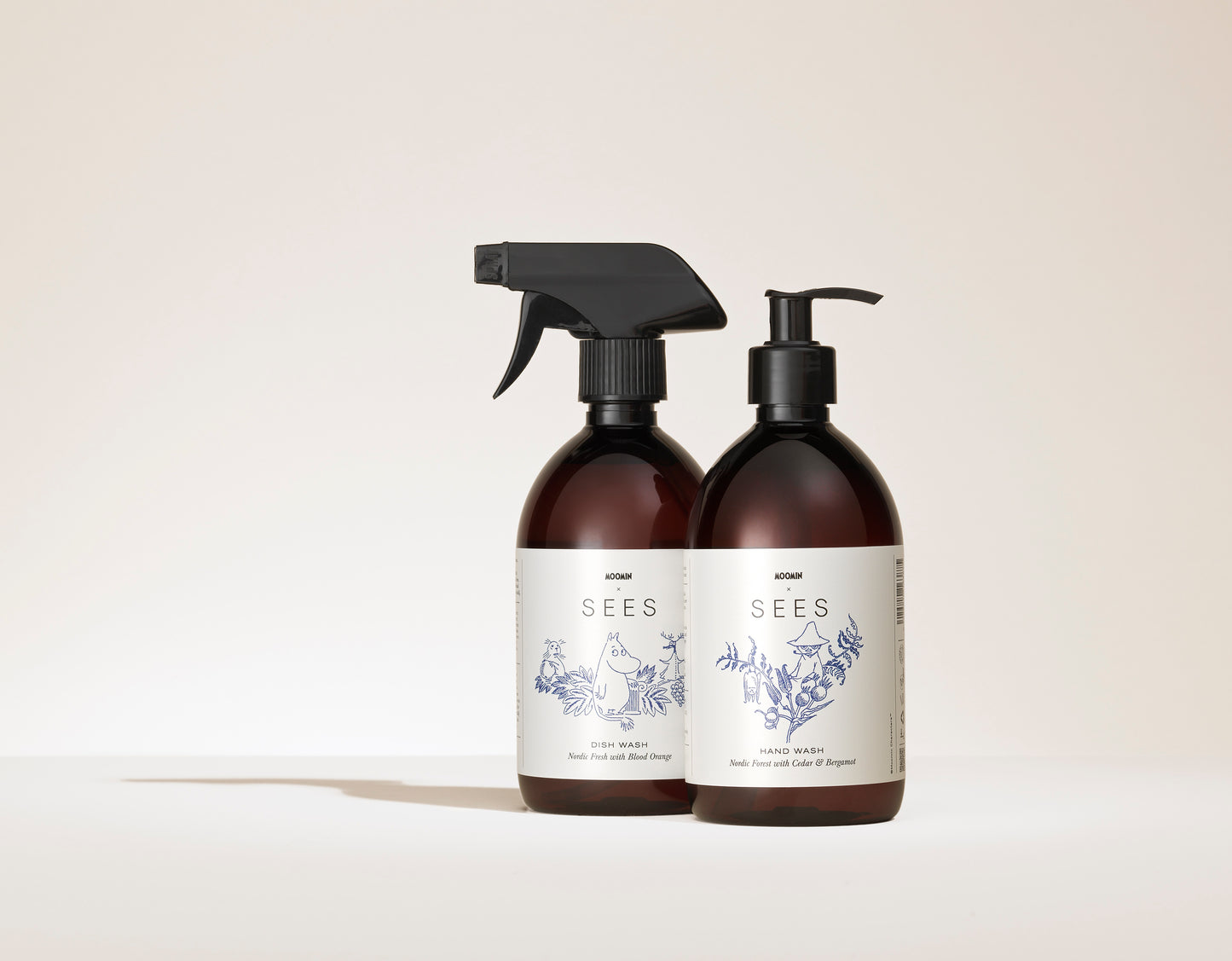 Due flaconi marroni con etichette bianche della SEES Company; uno è SEES x Moomin Dish Wash Spray con erogatore spray e detersivo biodegradabile per piatti, l'altro è Hand Wash con pompa. Entrambi presentano illustrazioni botaniche blu su sfondo neutro.