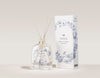 The SEES Company x Moomin Room Diffuser Nordic Summer, in una bottiglia di vetro trasparente con bastoncini di legno e una scatola bianca decorata con motivi floreali blu, cattura la fresca essenza dell'estate nordica su uno sfondo neutro.