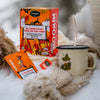 Avvolto in caldi abiti invernali, qualcuno versa una bevanda calda da un thermos in una tazza Snufkin accanto a una scatola e a pacchetti di tè rooibos Nordqvist All Things Fun Are Good For Your Tummy, il tutto disposto sulla neve.