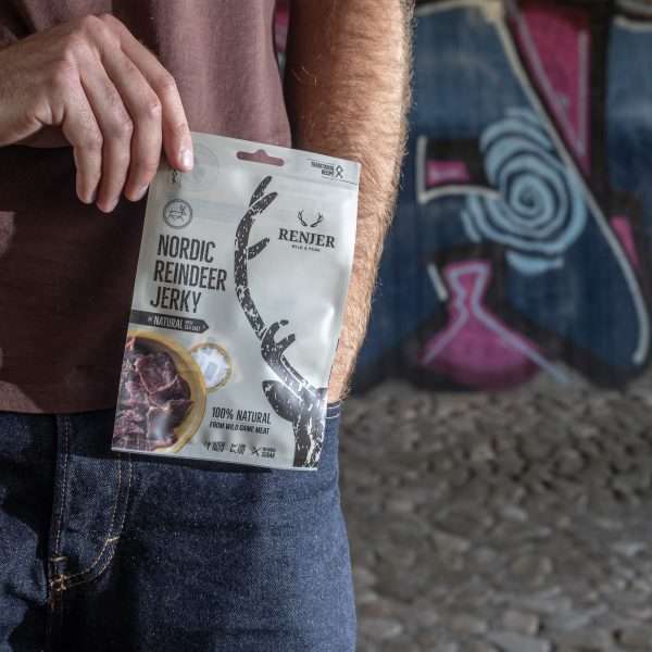 Qualcuno tiene in mano una confezione di Renjer Nordic Reindeer Jerky Sea Salt di Renjer davanti a un muro ricoperto di graffiti su un terreno roccioso, mettendo in risalto il sapore caratteristico della carne di renna della Lapponia in un ambiente urbano.