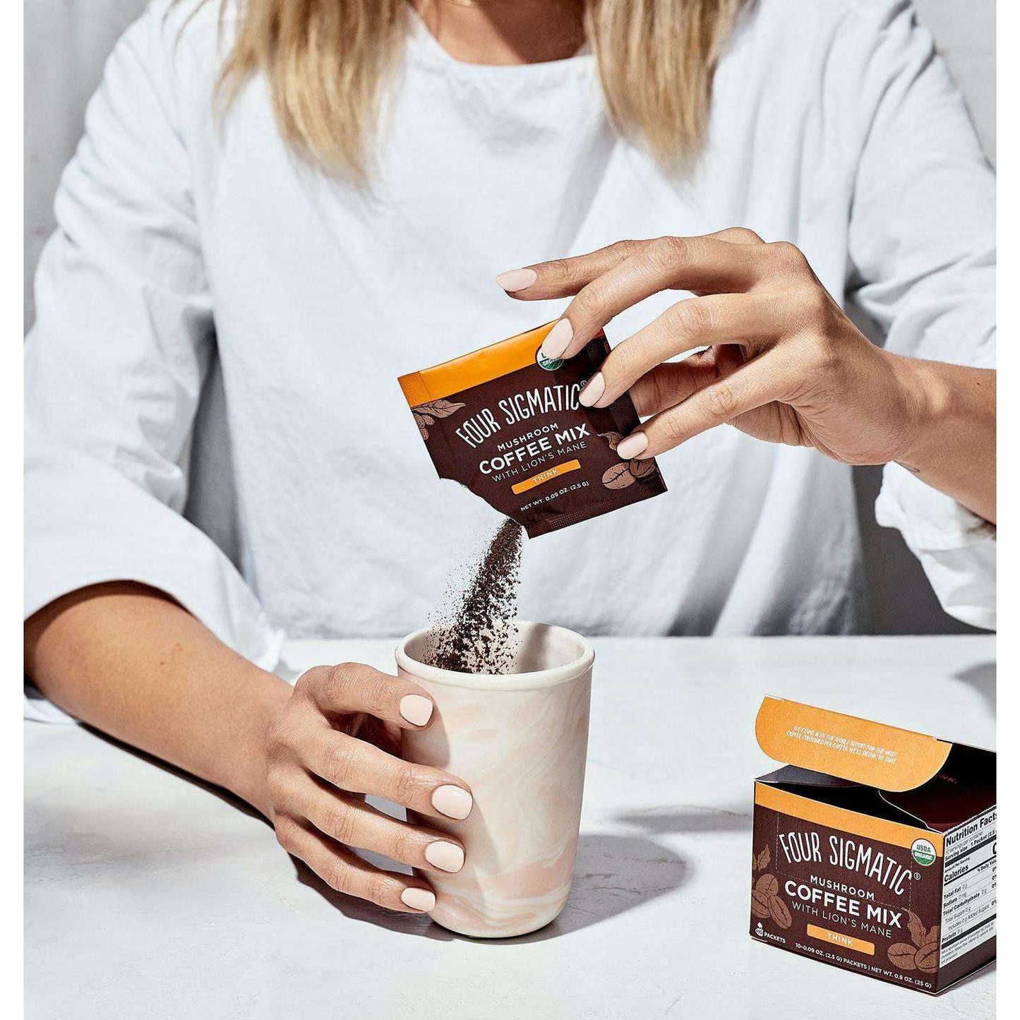 Una persona versa la polvere Four Sigmatic Mushroom Coffee Lions Mane da una bustina in una tazza di marmo, con la confezione Four Sigmatic visibile sul tavolo.
