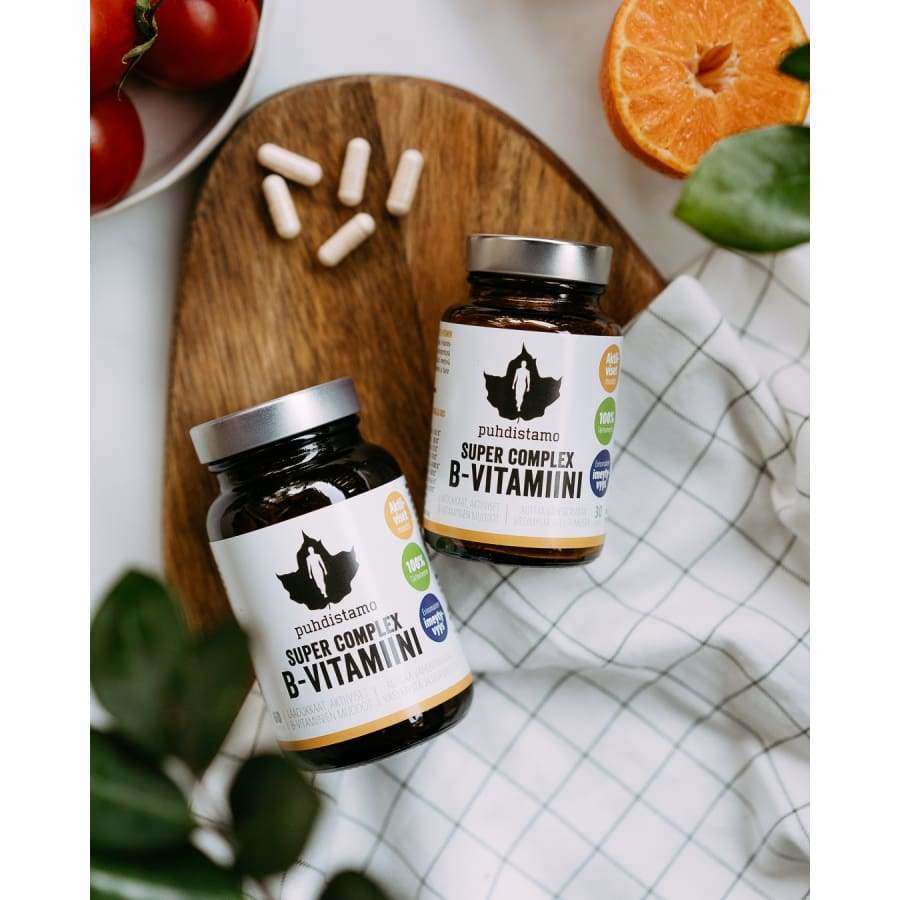 Due flaconi di Puhdistamo Super Complex B-complex di Puhdistamo sono appoggiati su una superficie bianca con un panno a quadretti, circondati da capsule, pomodori, una metà di arancia e foglie verdi.