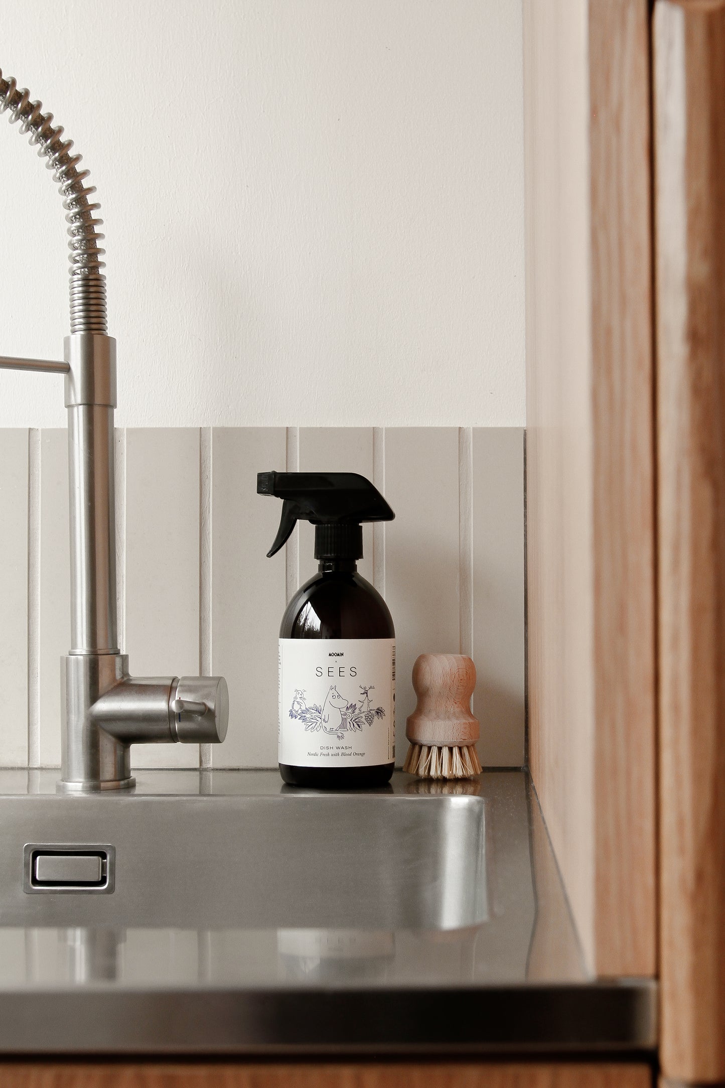 Een close-up van een aanrecht met een zilveren kraan, SEES x Moomin Dish Wash Spray SEES Companymet een veganistische formule en een kleine houten afwasborstel op het aanrecht tegen een betegelde achterwand.