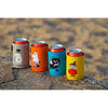 Vier blikjes frisdrank in Emendo Moomin Cooler Moominmamma neopreen hoesjes – grijs, oranje, blauw en geel – staan op een rots buiten, met Moomin-figuren zichtbaar tegen een wazige zandachtige achtergrond.