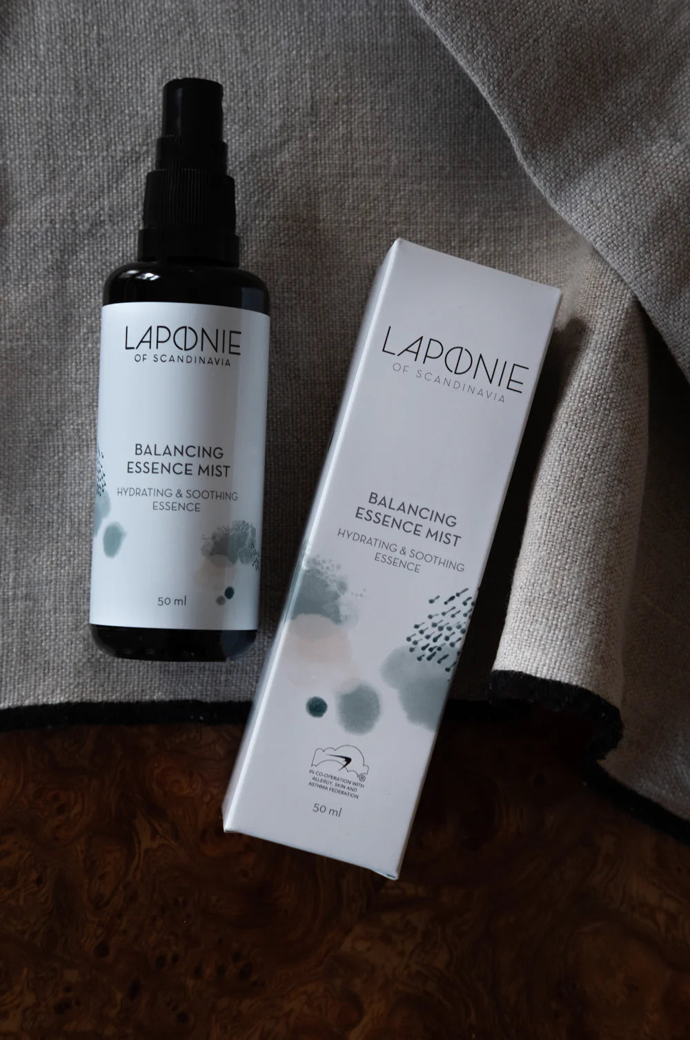 Een fles en doos Laponie of Scandinavia Essence Mist van Laponie of Scandinavia, een hydraterende toner en gezichtsmist voor de gevoelige huid, worden getoond op een grijze stoffen ondergrond met een bruine houten rand.