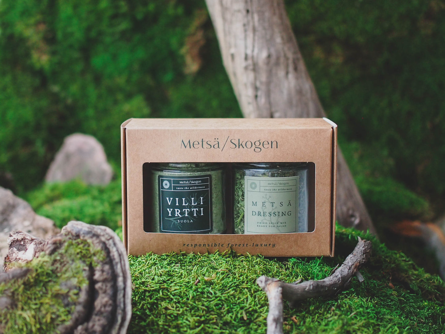 De Metsä/Skogen Forest Dressing & Wild Herb Salt Gift Set bestaat uit twee glazen potjes in een bruine Metsä/Skogen-doos met mos en hout, omgeven door groen. De potjes bevatten Finse wilde kruiden: Villi Yrtti-zout en Metsä-bosdressing.