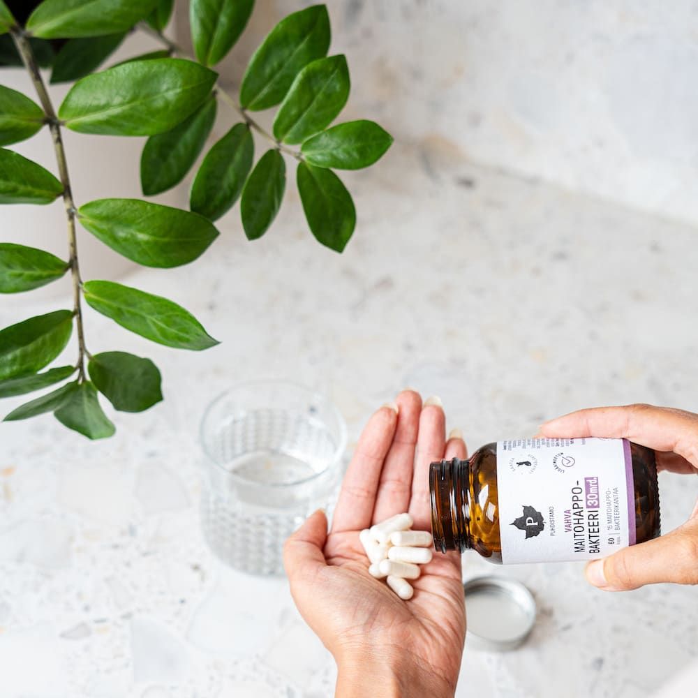 Een hand houdt witte capsules van Puhdistamo Strong Probiotic 30 miljard CFU vast, die uit een Puhdistamo-flesje zijn gegoten op een marmeren aanrecht naast een glas water en een groene plant met bladeren.