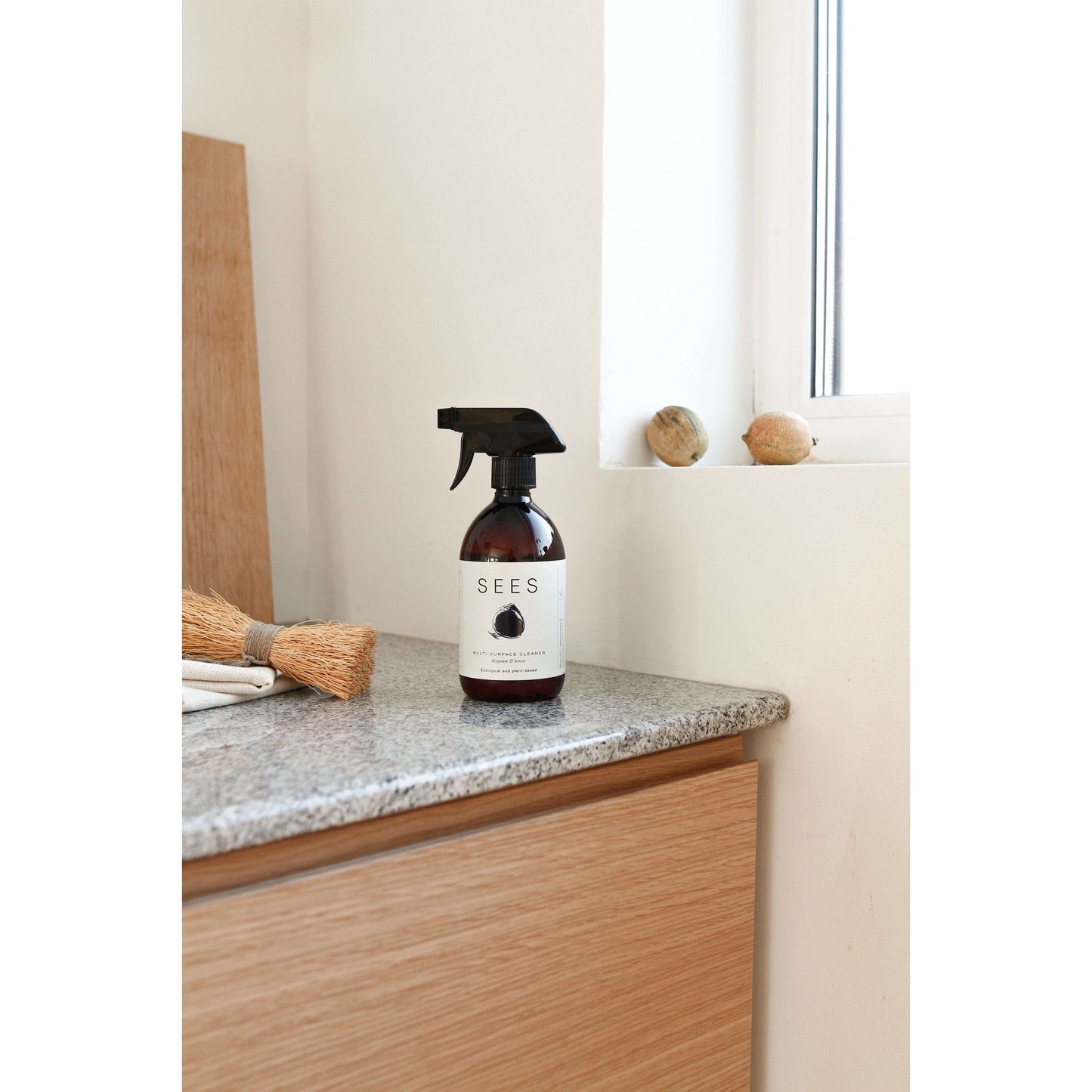 Een SEES Company voor de keuken en badkamer staat op een aanrecht bij het raam, naast een schoonmaakborstel en decoratieve stenen – ideaal voor iedereen die op zoek is naar een reinigingsspray op plantaardige basis.