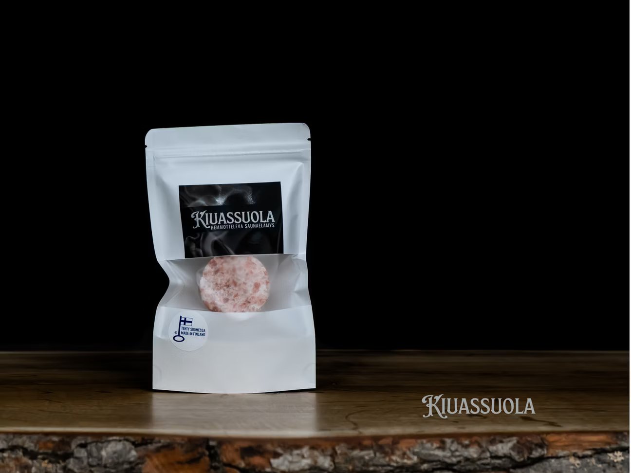 Een wit hersluitbaar zakje met het label "Sauna Salt" van het merk Kiuassuola bevat rond Himalaya-kristalzout. De verpakking ligt op hout tegen een zwarte achtergrond, met "Kiuassuola" in de rechterbenedenhoek.