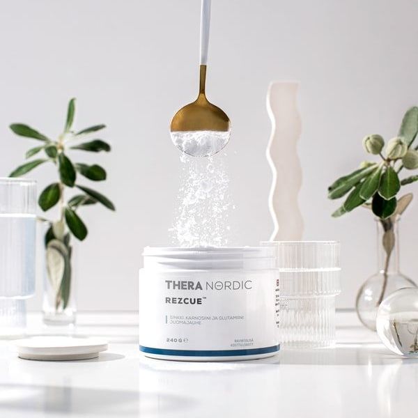 Een hand schept THERA Nordic Rezcue Drink Powder uit een lepel in een open bakje. De scène is helder, met glaswerk en groene planten op de achtergrond, waardoor dit product van THERA Nordic voor een gezonde darmflora extra goed uitkomt.