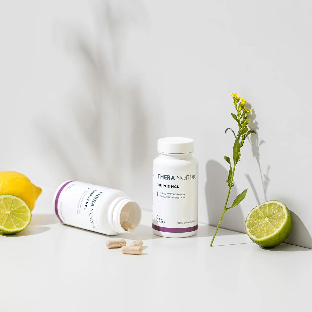 Twee THERA Nordic Triple HCL-flessen van THERA Nordic – één rechtopstaand en één omgekanteld met tabletten die eruit vallen – worden afgebeeld met een citroen, een gehalveerde limoen en een klein geel bloemetje, waardoor dit supplement voor een gezonde spijsvertering extra goed uitkomt tegen een lichte achtergrond.