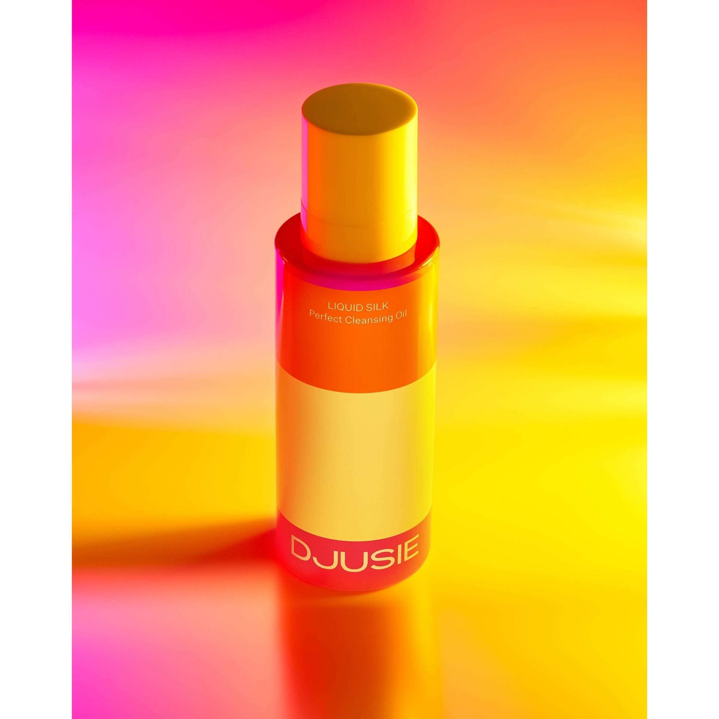 Een fles Djusie Liquid Silk Perfect Cleansing Oil staat rechtop tegen een levendige gele, oranje en roze kleurverloop. De strakke fles heeft een gele dop en opvallende witte branding, en verwijdert moeiteloos waterbestendige make-up.