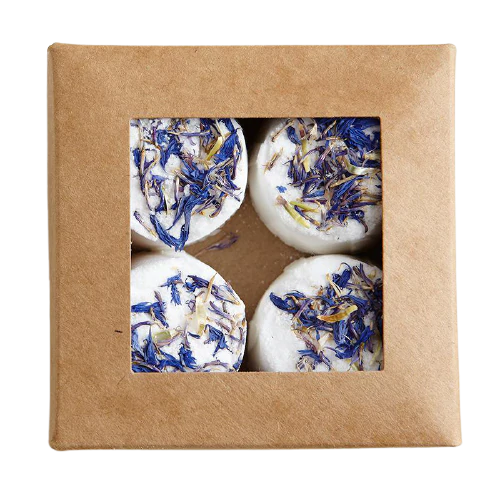 Een bruine kartonnen doos met een vierkant venster onthult vier ronde, witte Kaurilan Sauna Footbath-bommen van Kaurilan Sauna, bedekt met gedroogde blauwe en gele bloemblaadjes, handgemaakt in Helsinki van veganistische, natuurlijke ingrediënten.