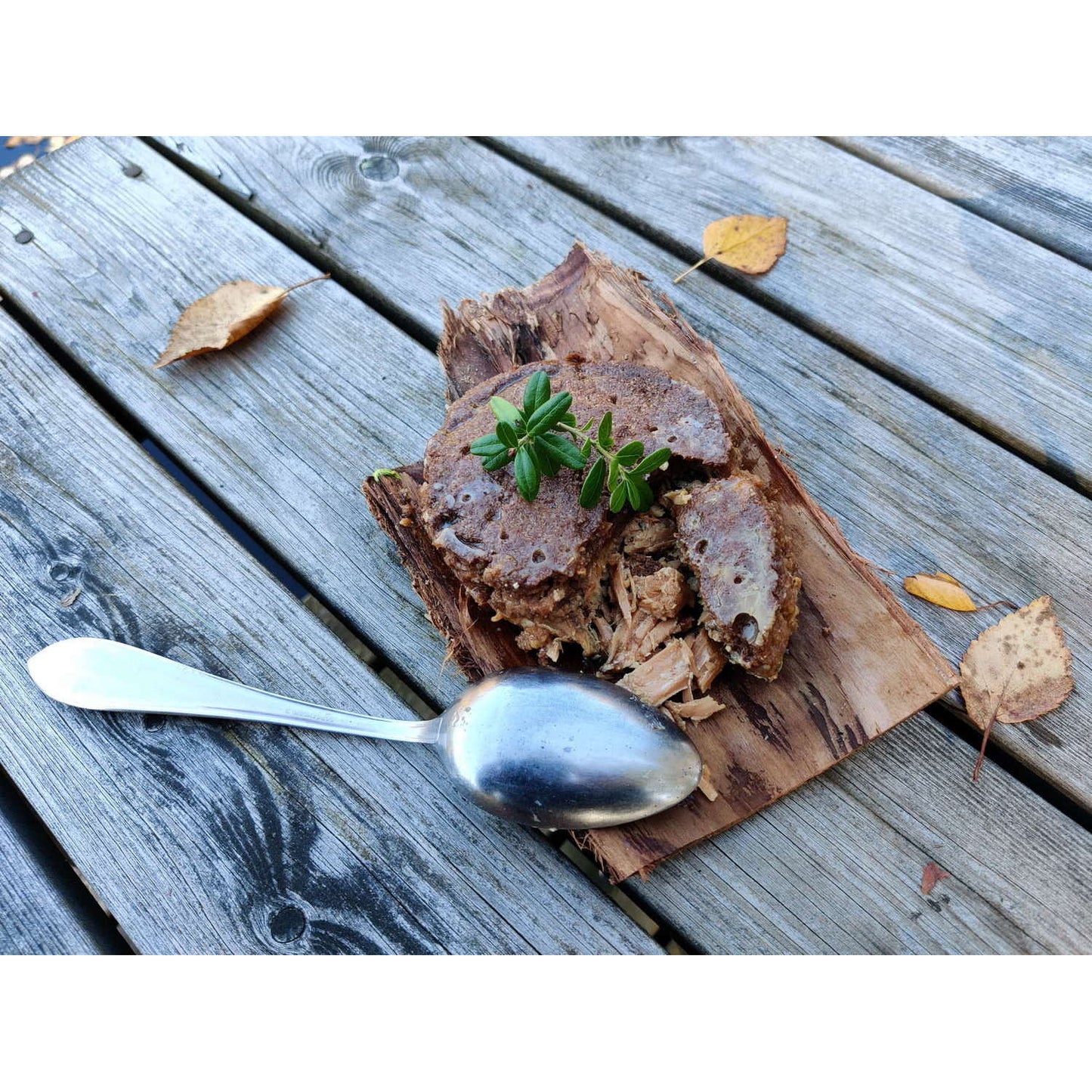 Een rustieke Finse lekkernij, Pielisen Kala Finnish Fish Pasty van Pielisen Kala, wordt geserveerd op boomschors met een zilveren lepel en herfstbladeren, waardoor de authentieke charme van traditioneel Muikkukukko-flatbread en vis goed tot zijn recht komt.