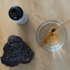 Een pipet met amberkleurig biologisch chaga-extract van KÄÄPÄ Mushrooms boven een glas water. Vlakbij staan Finse chaga en de open fles met extract op een licht houten oppervlak.