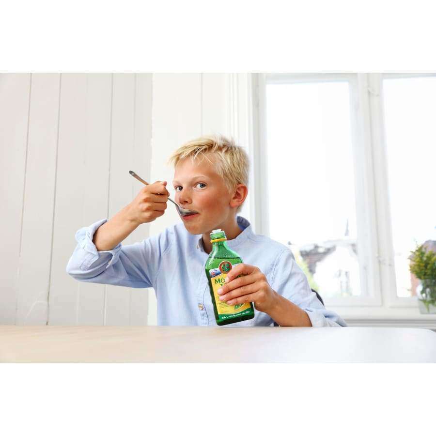 Een jonge blonde jongen in een lichtblauw shirt zit binnen aan een tafel en eet een lepel Möller-levertraan van Möller, met witte muren en een raam op de achtergrond.