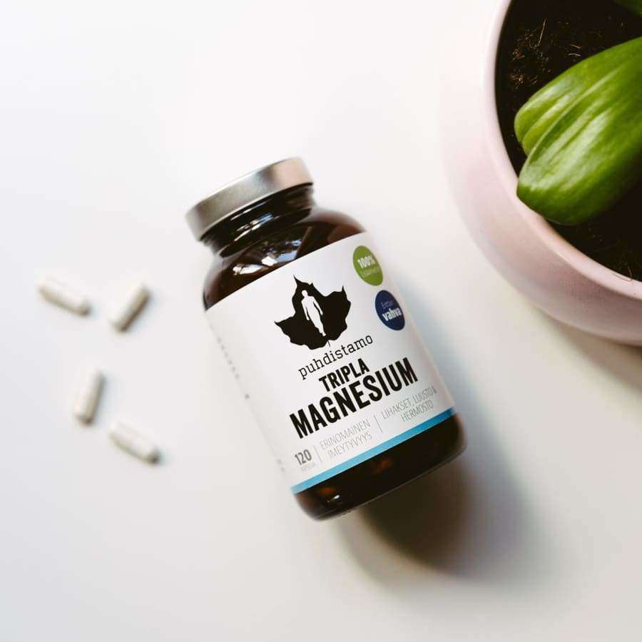 Een fles Puhdistamo Triple Magnesium van Puhdistamo staat op een wit oppervlak naast drie witte capsules en een groene potplant, waarmee dit goed opneembare supplement wordt gepresenteerd dat ideaal is voor het ondersteunen van de gezondheid van het hart.